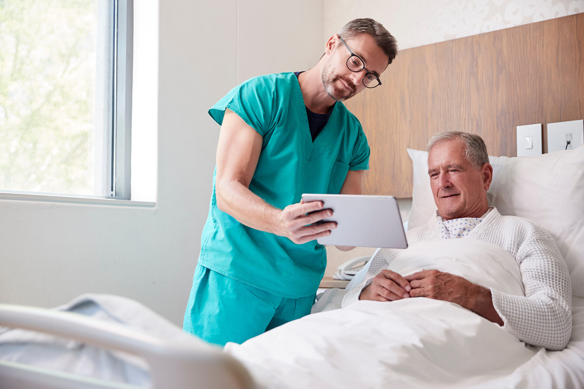 Älterer Mann liegt im Krankenhausbett, während ein Arzt in grüner OP-Kleidung ihm etwas auf einem Tablet zeigt. Darstellung von medizinischer Versorgung, Beratung und Leistungen einer Krankenzusatzversicherung.