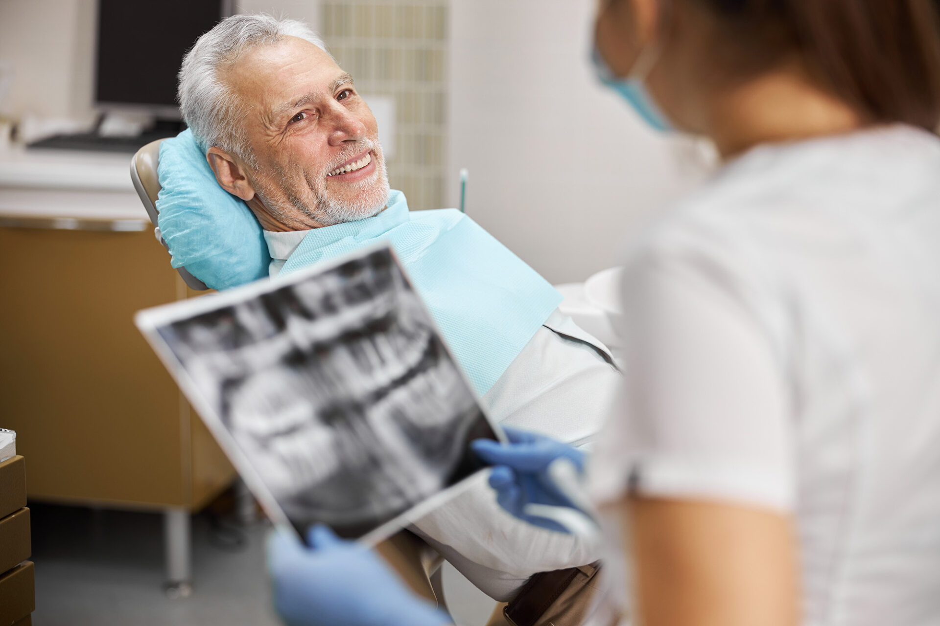 Älterer Mann sitzt lächelnd auf dem Zahnarztstuhl, während eine Zahnärztin ihm ein Röntgenbild seiner Zähne zeigt. Darstellung von professioneller Zahnbehandlung und Patientenbetreuung.