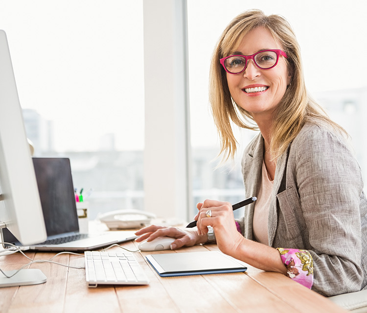 Frau mit Brille sitzt am Schreibtisch. Sie hält einen Stift in der linken Hand und die Computer-Maus in der rechten.