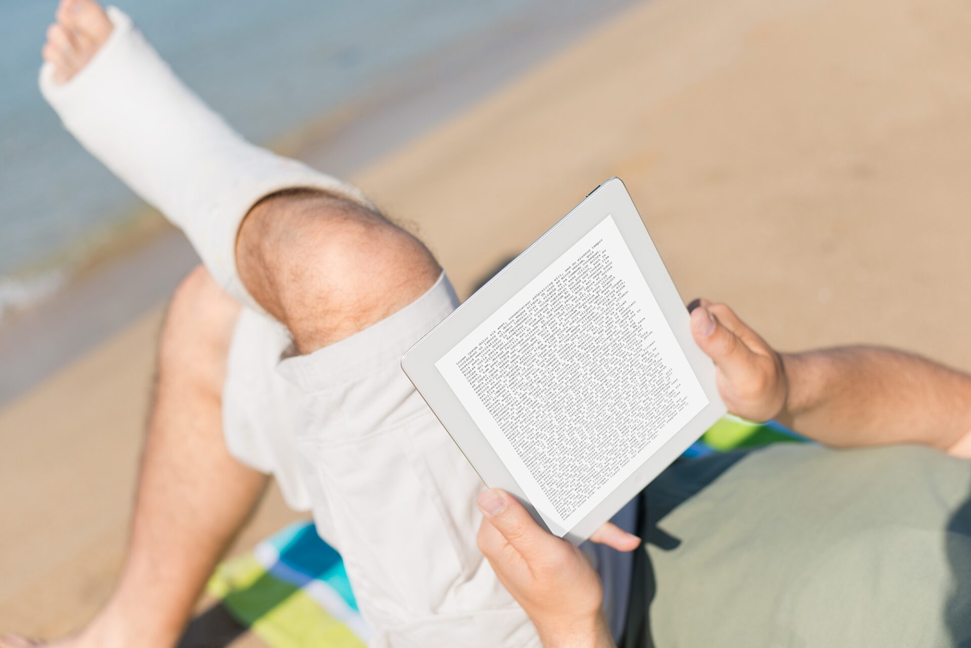 Mann mit Gips am Fuß sitzt mit verschränkten Beinen am Strand und liest ein Buch auf einem Tablet.