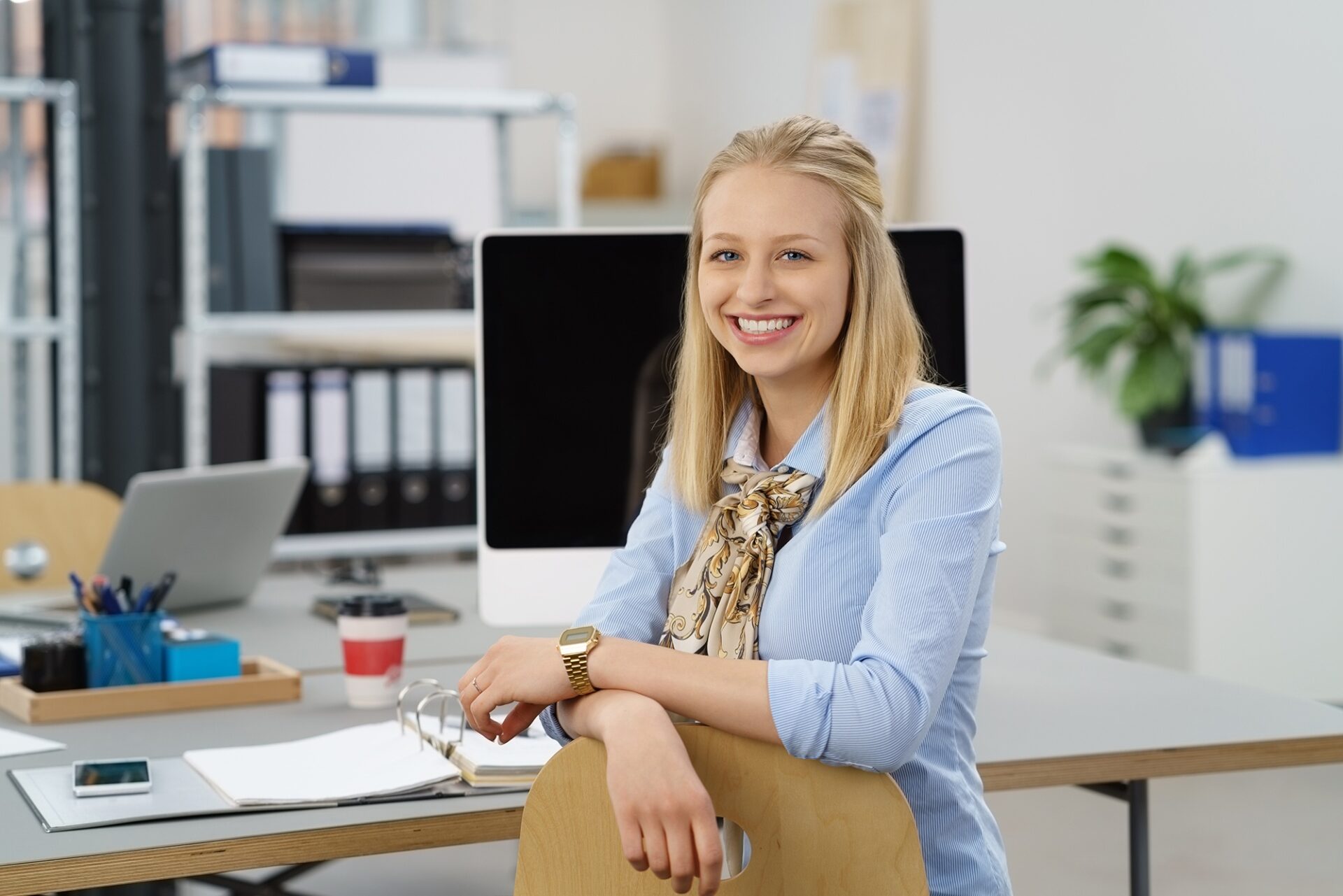 Junge Frau sitzt lächelnd in einem modernen Büro vor einem Computer und Ordnern. Darstellung von Professionalität, Beratung und betrieblicher Altersvorsorge.