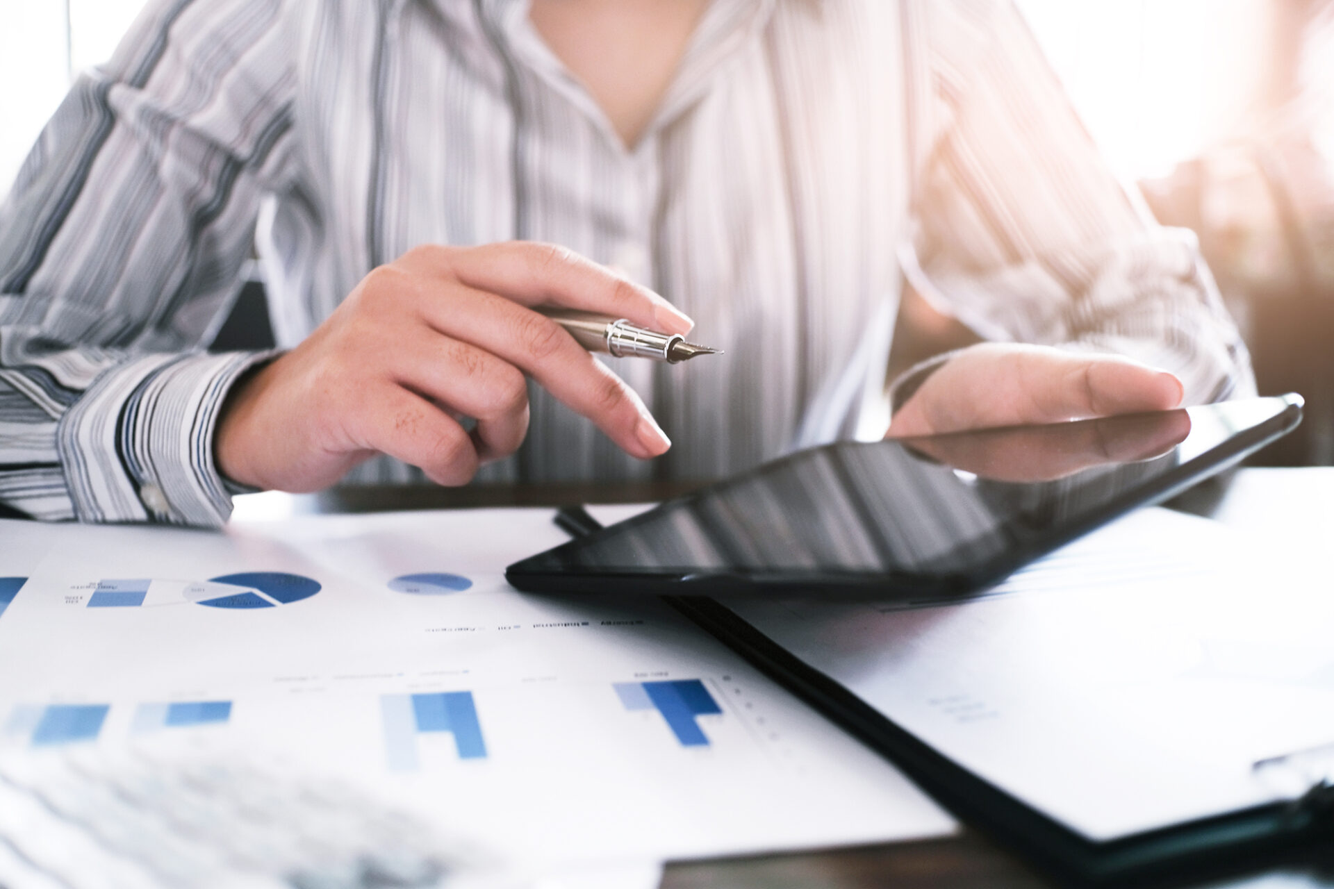 Nahaufnahme einer Person in Businesskleidung, die mit einem Stift auf ein Tablet zeigt und Diagramme auf Papier vor sich liegen hat. Symbolisiert Analyse, Dokumentation und digitale Prozesse im Rahmen der Compliance- und Beratungsarbeit von S mobile.