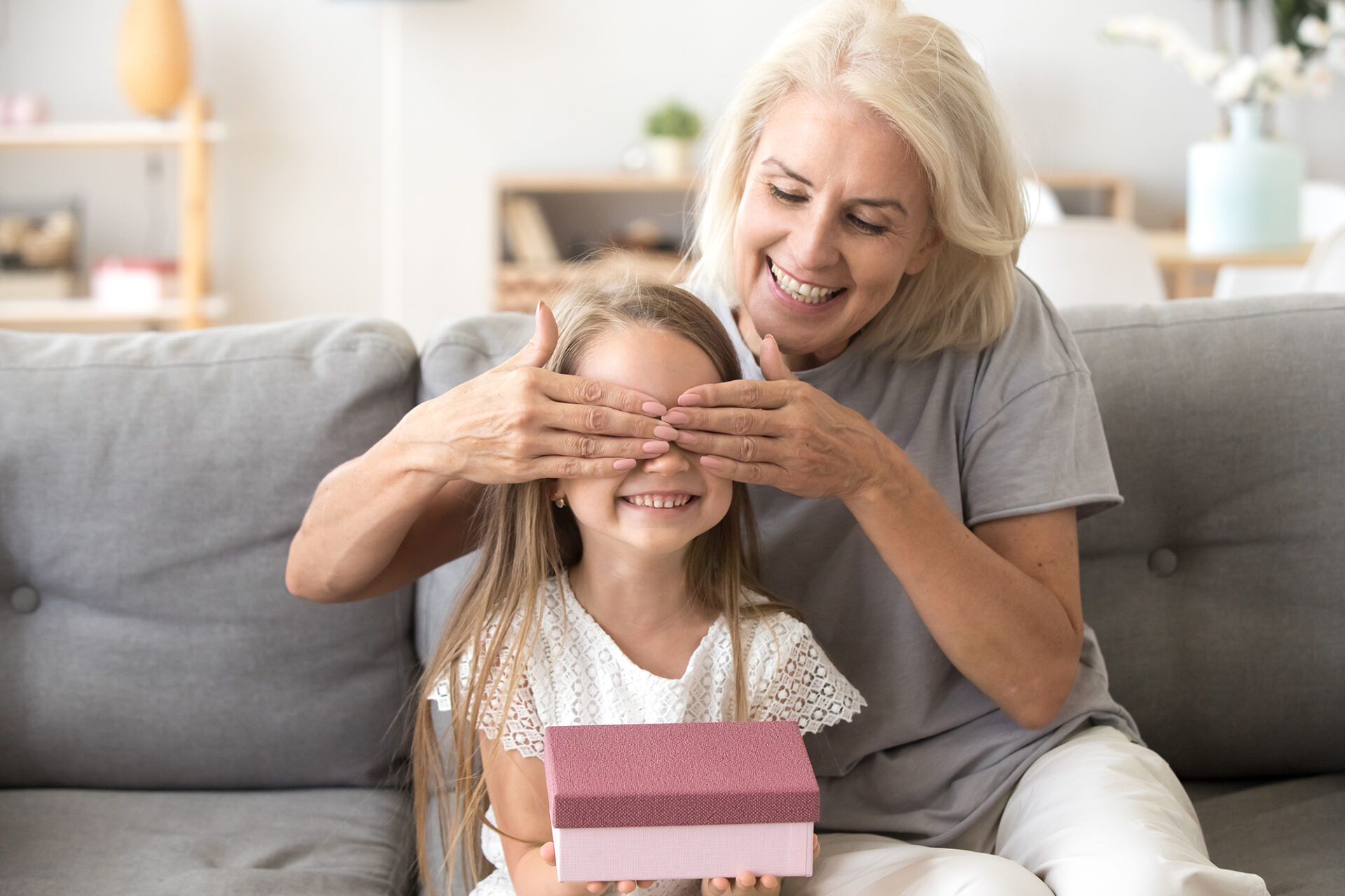 Ältere Frau sitzt mit einem Mädchen auf dem Sofa und hält dem Kind spielerisch die Augen zu. Das Mädchen lächelt und hält eine kleine Geschenkbox in den Händen. Symbol für Fürsorge, Überraschungen und Generationenverbundenheit.