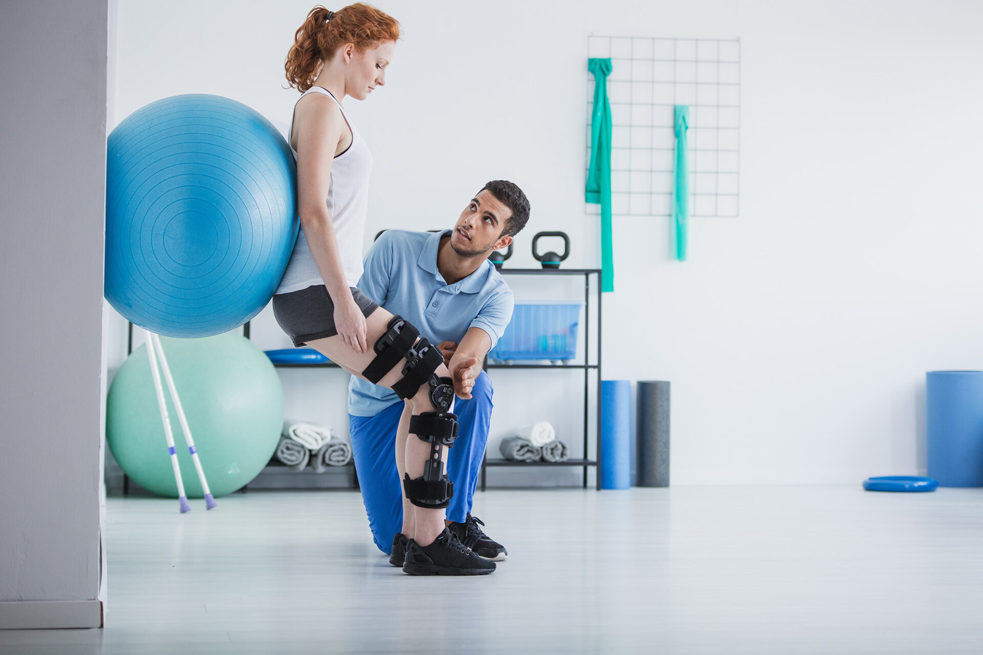 Eine Frau mit Knie-Orthese bei der Physiotherapie. EIn Therapeut begleitet sie bei einer Übung in einem Trainingsraum.