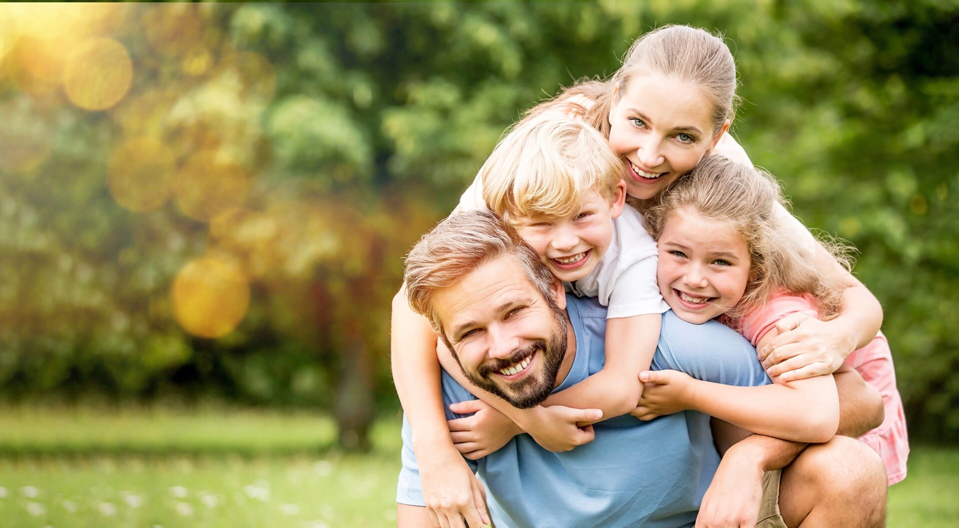 Eine Familie bestehend aus Vater, Mutter und zwei Kindern vor einem grünen Garten. Der Vater buckelt die beiden Kinder und wird von der Mutter umarmt.