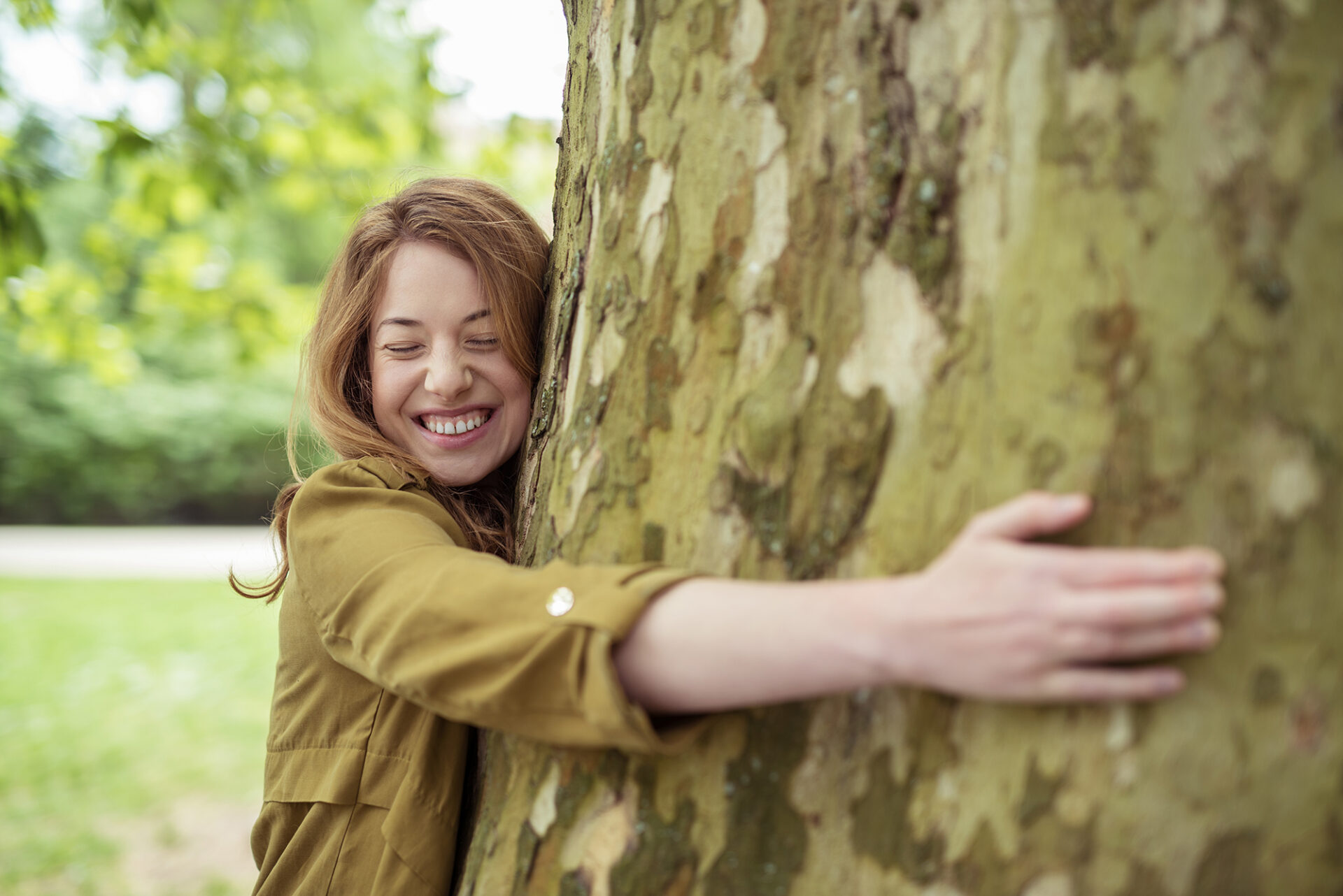 Junge Frau mit geschlossen Augen umarmt lächelnd einen Baum in einer grünen Parklandschaft. Symbol für Umweltbewusstsein, Naturverbundenheit und das Nachhaltigkeitsengagement der Firma S mobile.