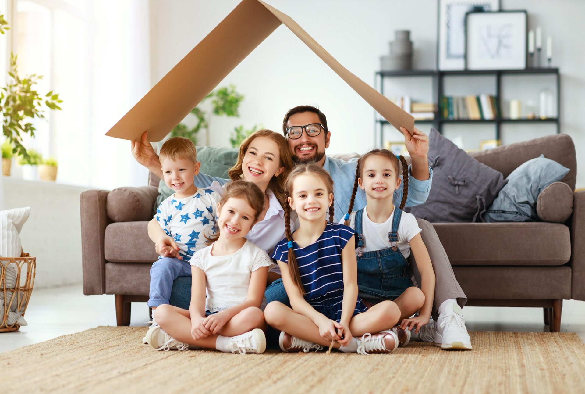 Glückliche Familie mit drei Kindern sitzt lachend im Wohnzimmer. Die Eltern halten ein großes Stück Pappe schützend wie ein Dach über die Familie. Symbol für Sicherheit, Geborgenheit und Vorsorge – passend zur Riester-Rente.
